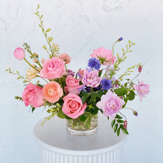 A floral arrangement with pink, lavendar, and peach flowers in a short glass vase